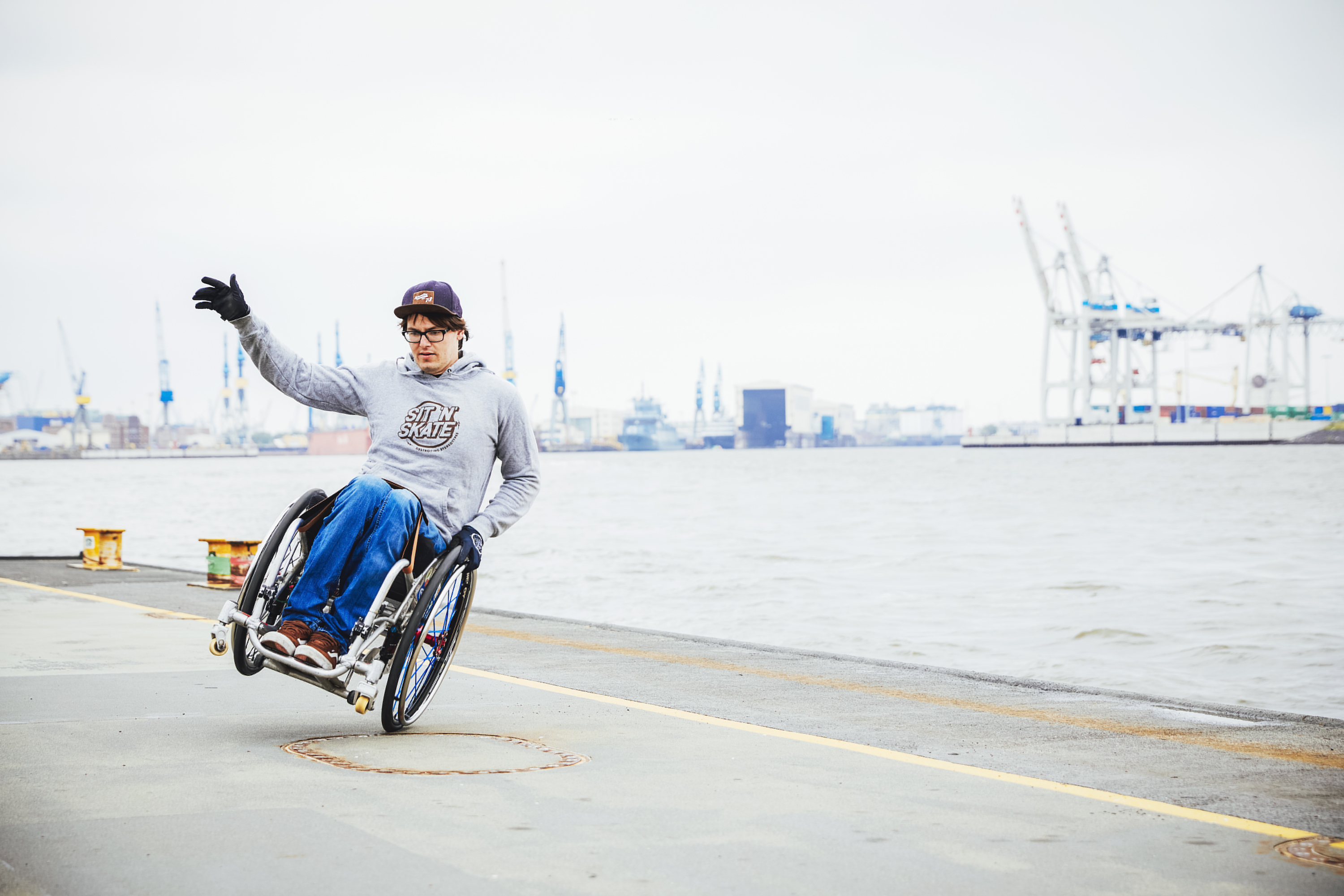 Ein Bild von Rollstuhlskater David Lebuser beim Skaten mit seinem Rollstuhl an der Hafenkante. Im Hintergrund sind Schiffe zu sehen..